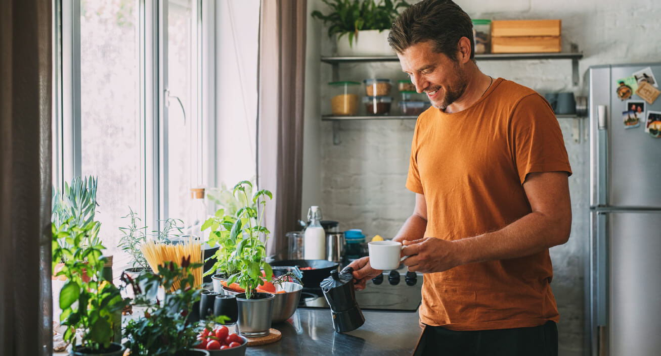 A man holds his coffee mug and coffee maker while smiling, indicating he just brewed coffee at home in order to develop better money habits.
